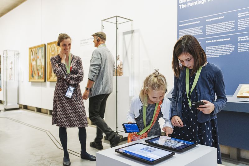 Besucherinnen an Tablets auf einem weißen Pult, im Hintergrund Gemälde und eine Vitrine, LVR-LandesMuseum Bonn