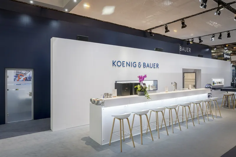 Lange weiße Theke mit Barhockern vor der Koenig-&-Bauer-Wand, darüber Strahler an der Traverse