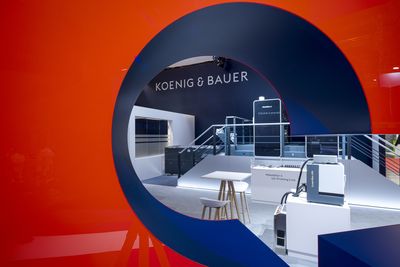 Blick durch eine runde Öffnung in einer roten Wand auf den Koenig-&-Bauer-Stand mit Logo, Theke, Barhockern und Maschinen