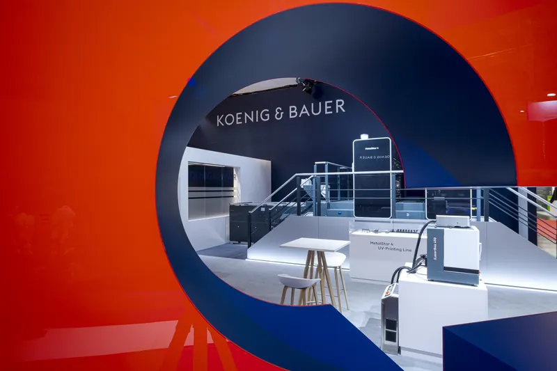 Blick durch eine runde Öffnung in einer roten Wand auf den Koenig-&-Bauer-Stand mit Logo, Theke, Barhockern und Maschinen