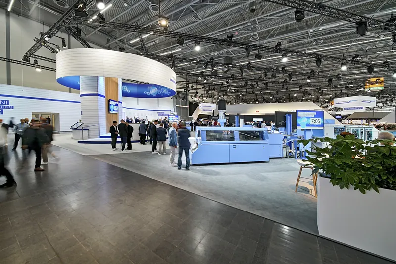 Maschinenreihe am Müller-Martini-Stand mit Besuchern, links der Turm, rechts Pflanzen
