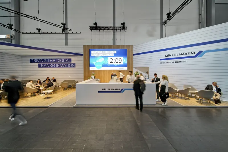 Empfangstheke am Müller-Martini-Stand vor einer Holzwand mit großem Bildschirm, davor Sitzgruppen